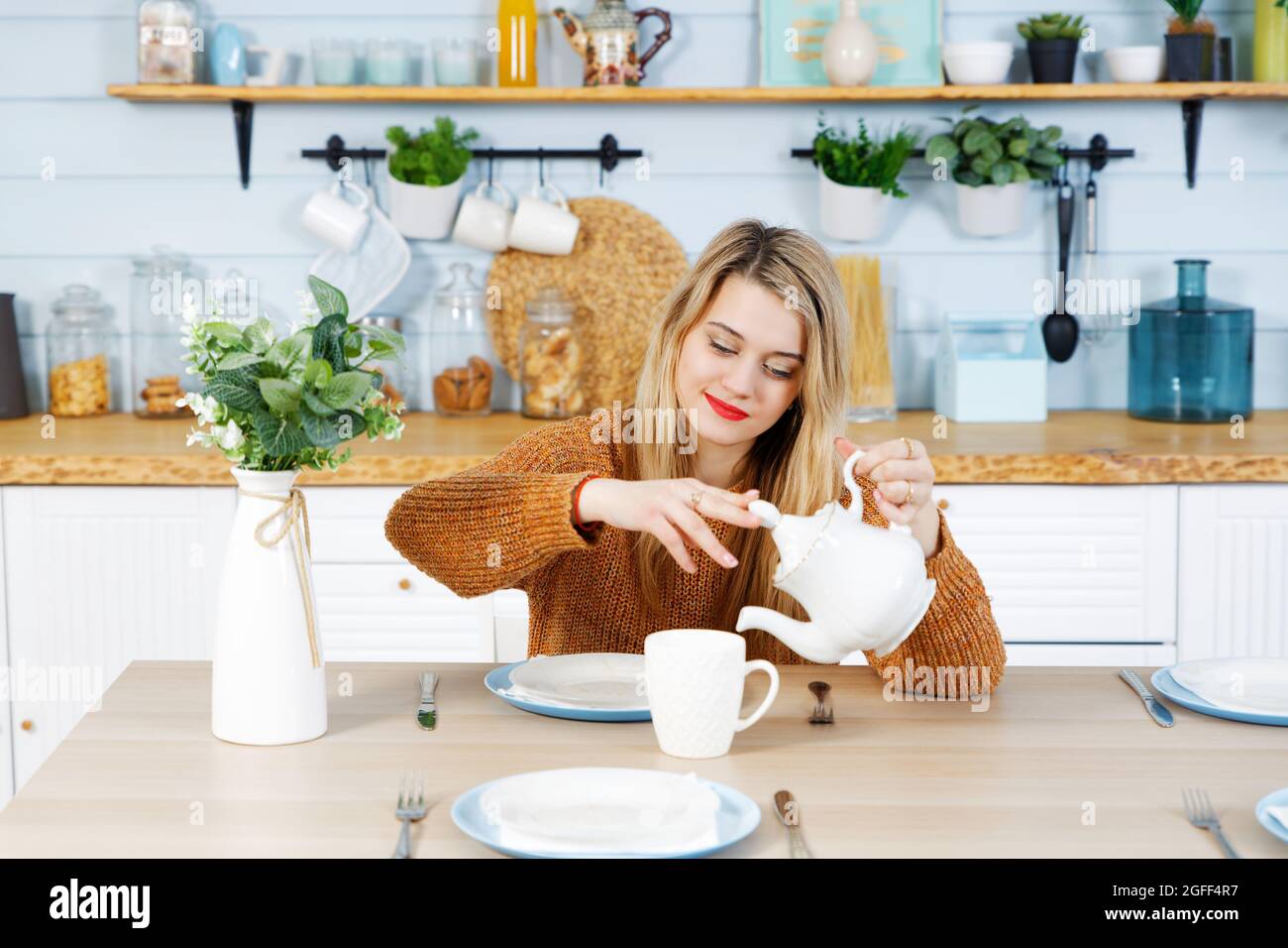 Attractive girl pours tea from a white teapot into a cup while sitting at the table in the domestic kitchen Stock Photo