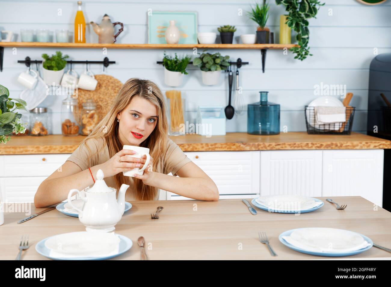 Young woman sitting at the kitchen table with a mug in her hands. Blurred background. Stock Photo