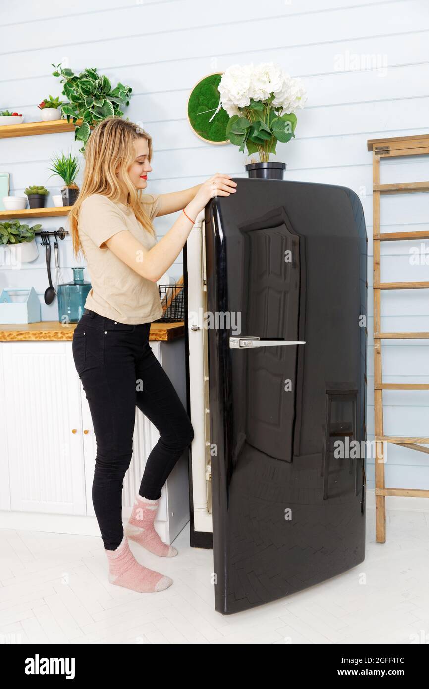 Young pretty woman open a door of a black refrigerator on the domestic kitchen. Stock Photo