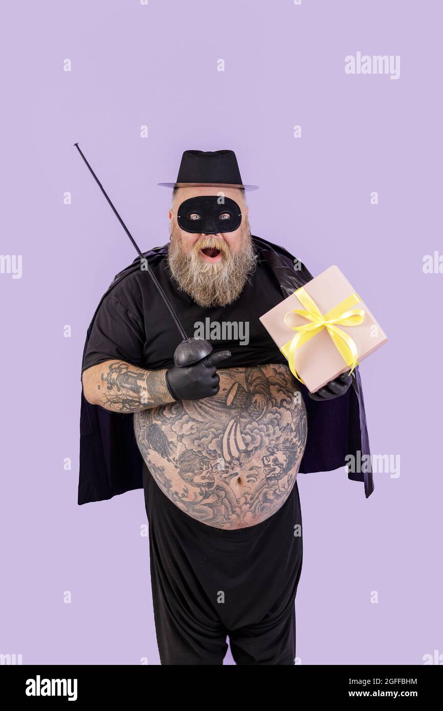 Surprised mature man with overweight in Zorro suit points onto gift box on purple background Stock Photo