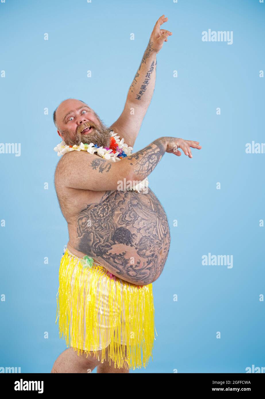 Funny obese man in decorative grass skirt and garland dances on light blue background Stock Photo