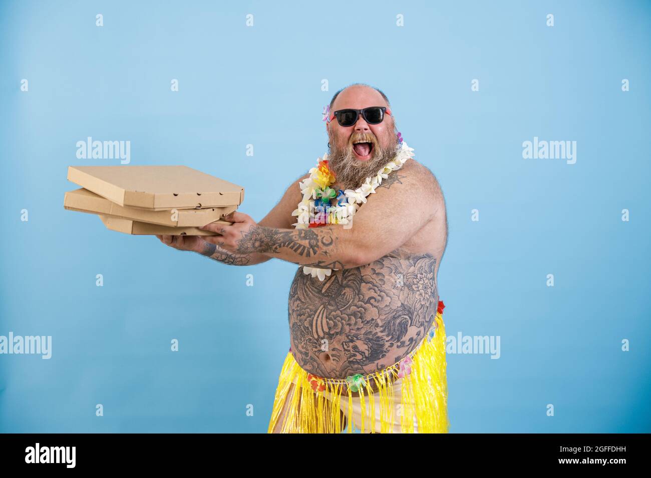 Happy obese man in grass skirt and flowers garland holds pizzas on light blue background Stock Photo