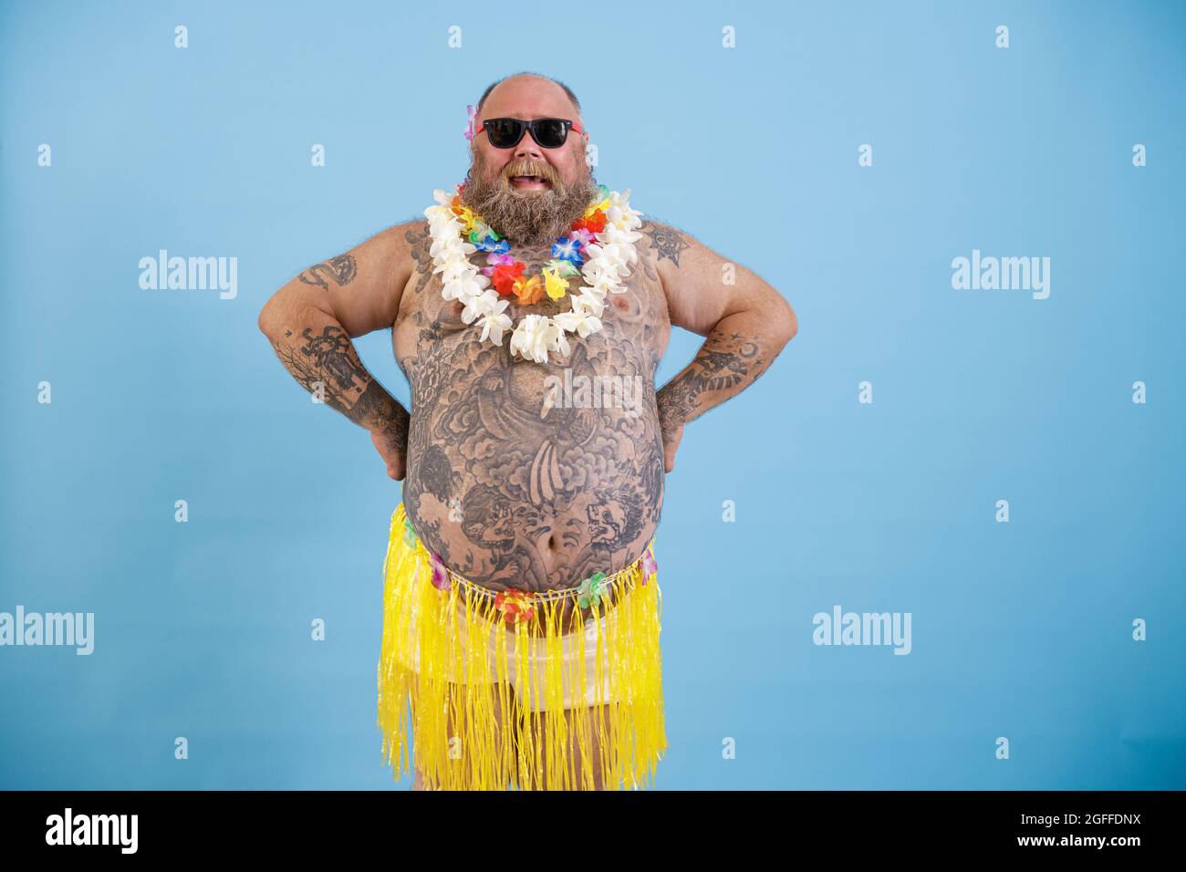 Happy man with overweight and tattooed tummy in decorative grass skirt on light blue background Stock Photo