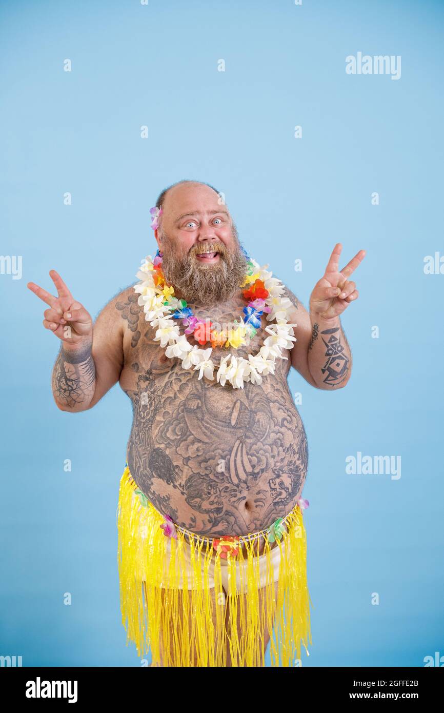 Cheerful obese man in decorative yellow grass skirt shows peace gestures on light blue background Stock Photo Alamy