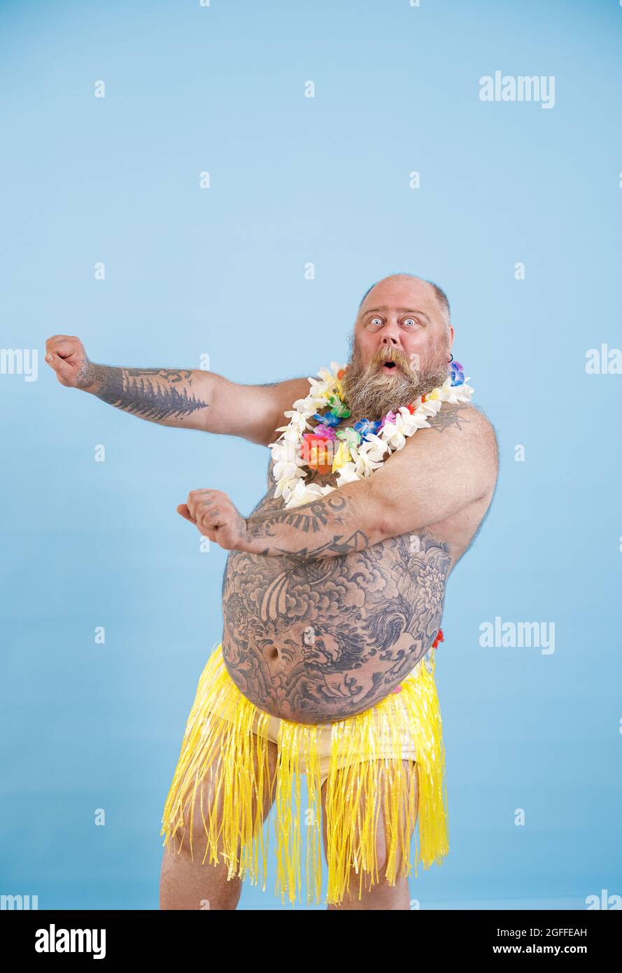 Emotional man with overweight in decorative grass skirt adances on light blue background Stock Photo