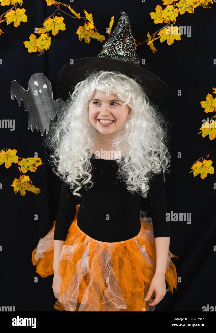 A cheerful girl in a white wig and a witch costume on a black background with ghosts. Funny Horror. A glance to the side Stock Photo