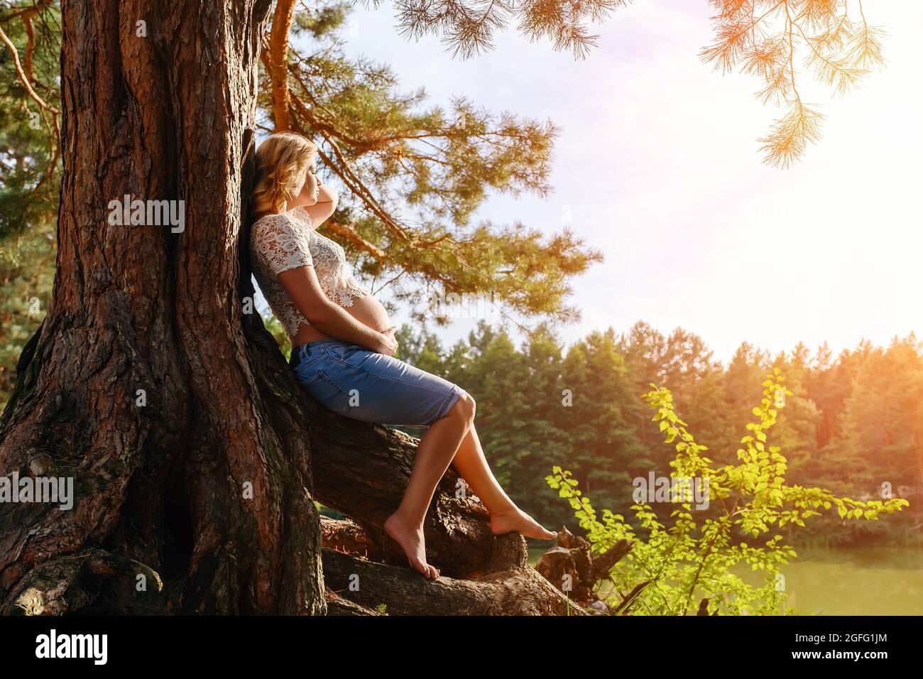 Young pregnant woman sits on the roots under a tree by the river Stock Photo