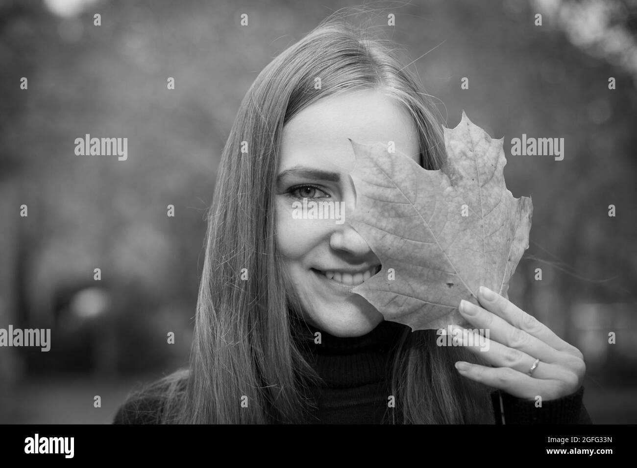 Beautiful young woman on a background of autumn trees. Emotion concept. The girl holds a maple leaf in her hand and covers one eye with the leaf. Stock Photo