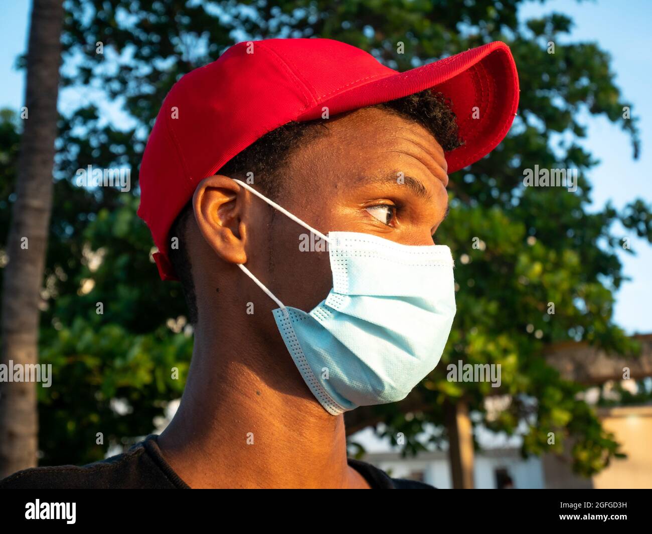 Riohacha, La Guajira, Colombia - May 26 2021: Portrait of a Young Latin Man with a Red Cap and a Protective Mask is  Looking at the Sunset with Trees Stock Photo