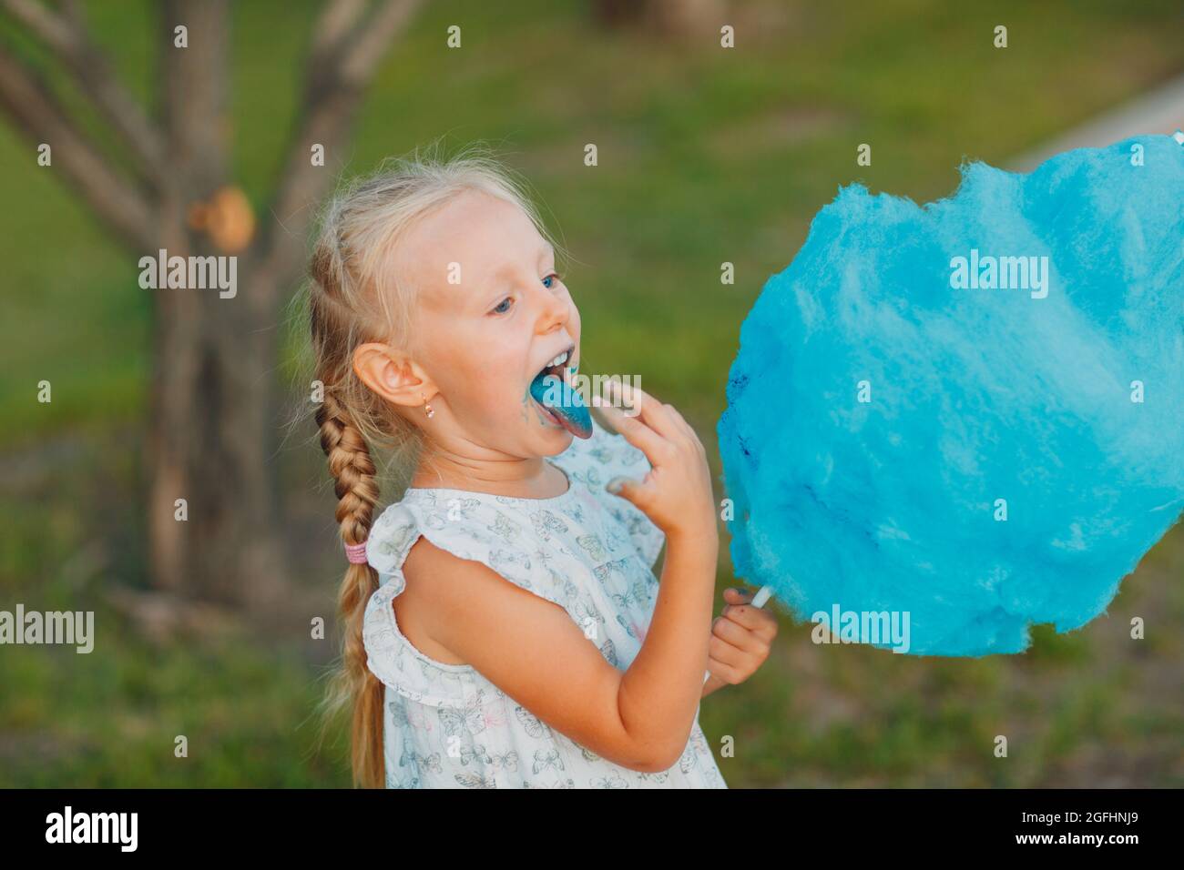 Little blond girl eating cotton candy and shows blue tongue in the park