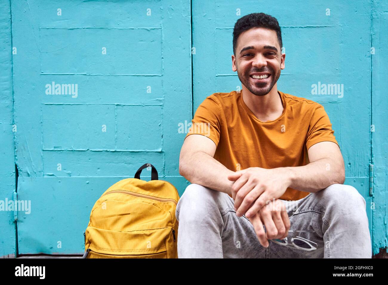 Happy young latino man sitting looking at camera with a backpack on one side. Resting from a walk in the city. High quality photo Stock Photo
