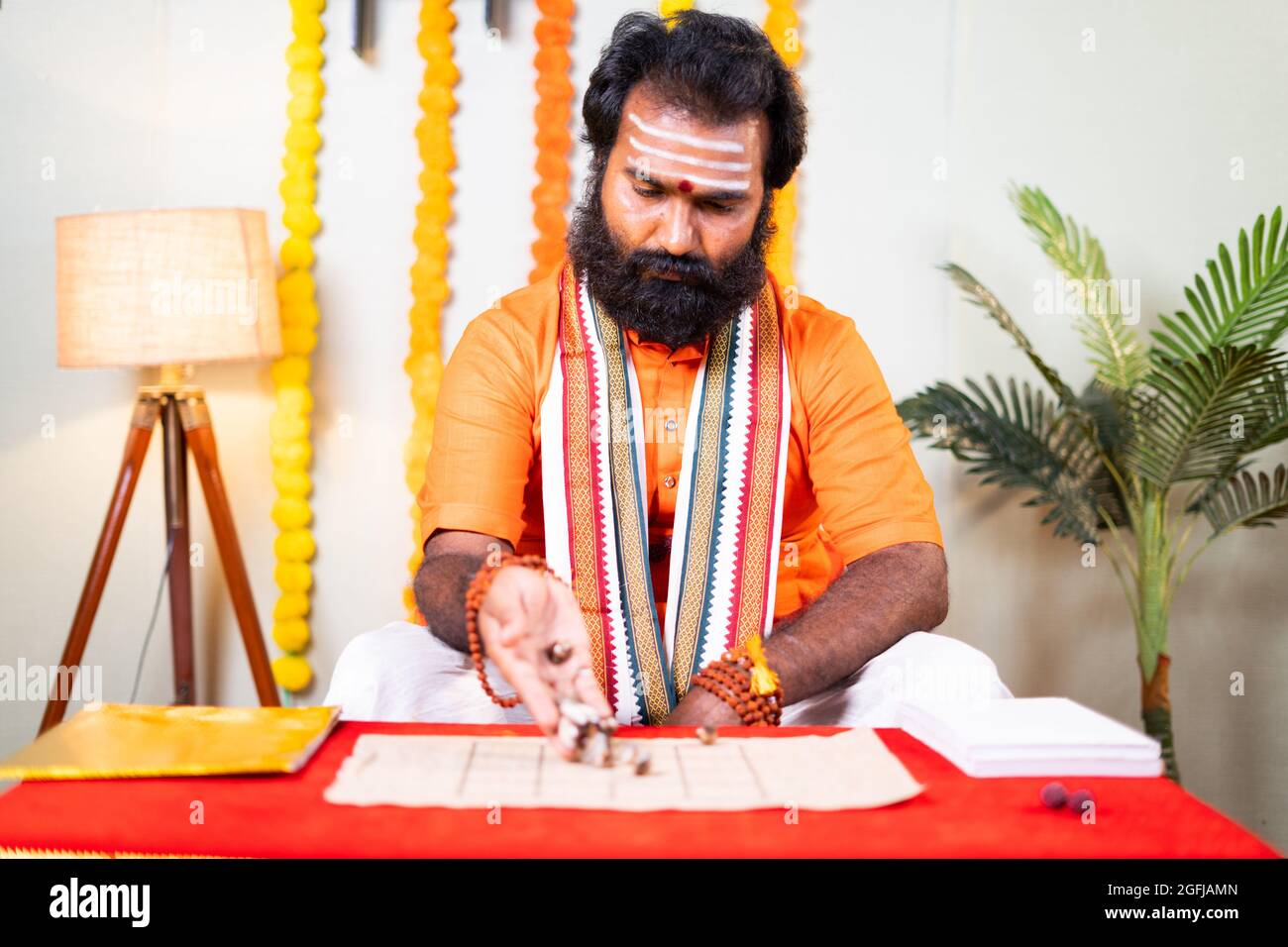 Beard priest, holy astrologer guru or jyotishi placing cowrie shells on chart and counting to predict future by looking into book - Concept of Indian Stock Photo