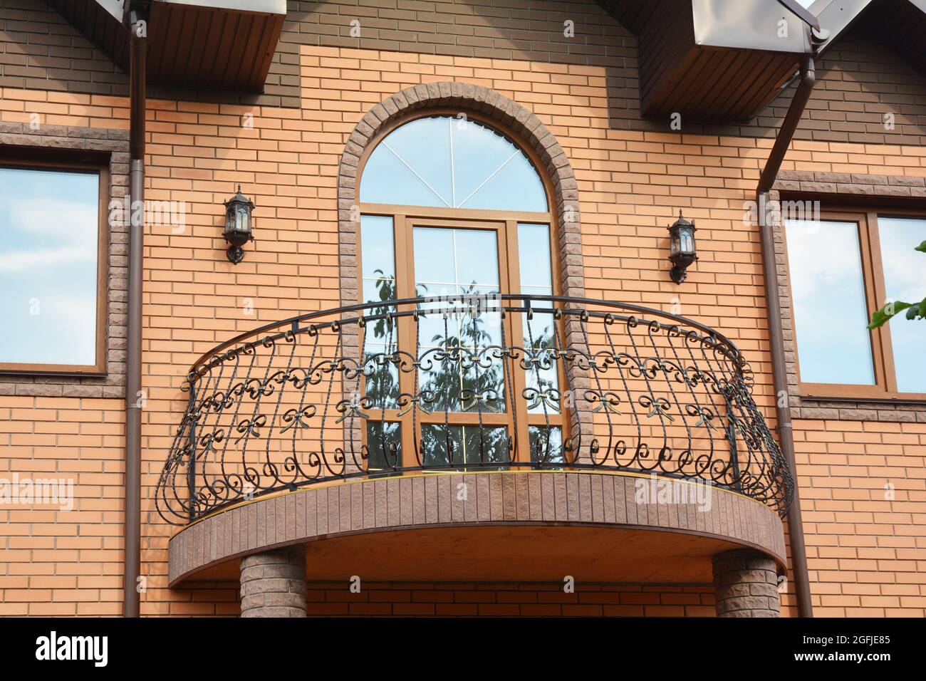 A close-up on semi-circled balcony with wrought iron railings, arched glass door and roof gutter system with downpipes of a brick house. Stock Photo