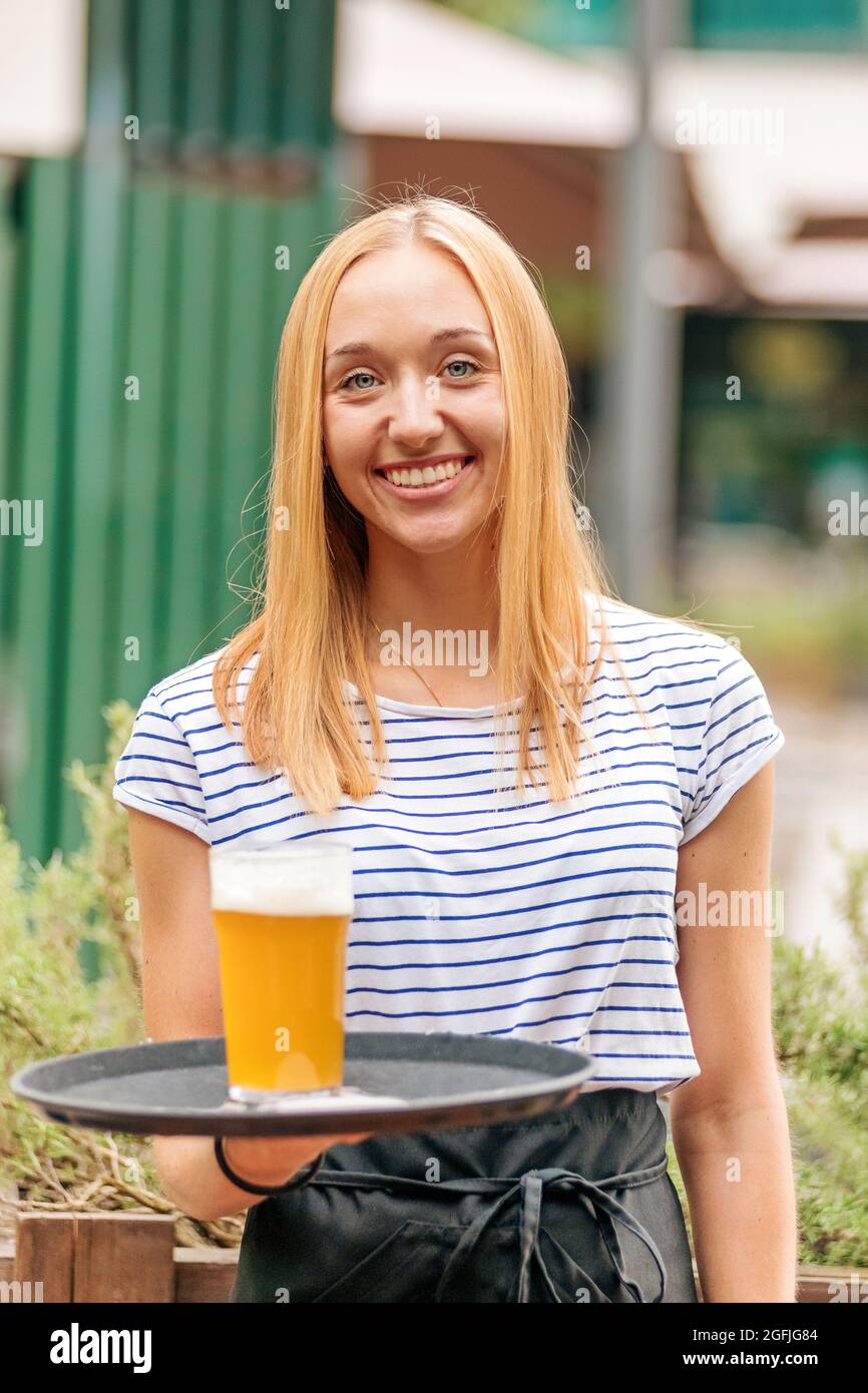 Happy friendly young female waitress delivering a glass of cold beer on a tray with a beaming smile at an outdoor restaurant or pub Stock Photo