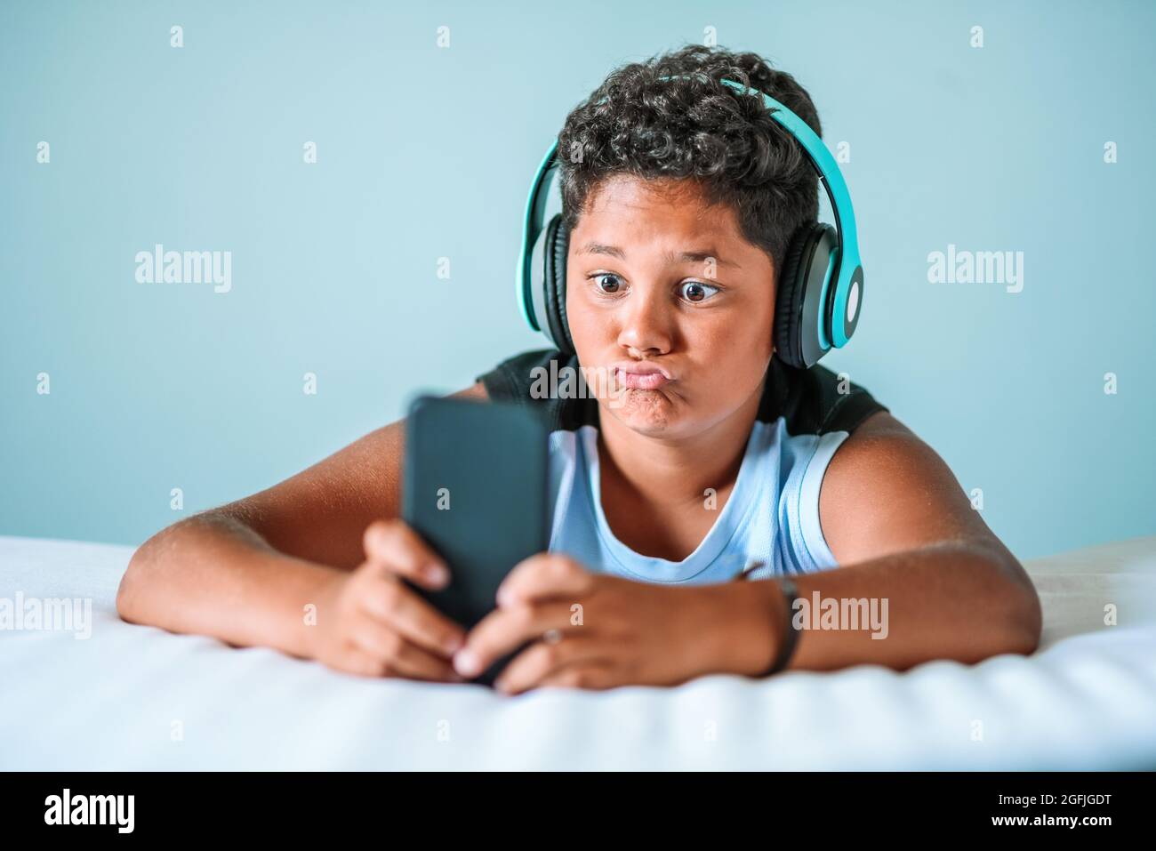 Charismatic young boy pulling a quirky face while listening to music on stereo headphones using his mobile phone Stock Photo