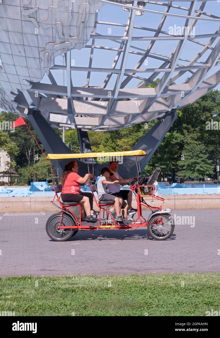 A family of four ride a rented Wheel Fun 4 wheel surrey cycle past the Unisphere in Flushing Meadows Corona Park in Queens, New York City. Stock Photo