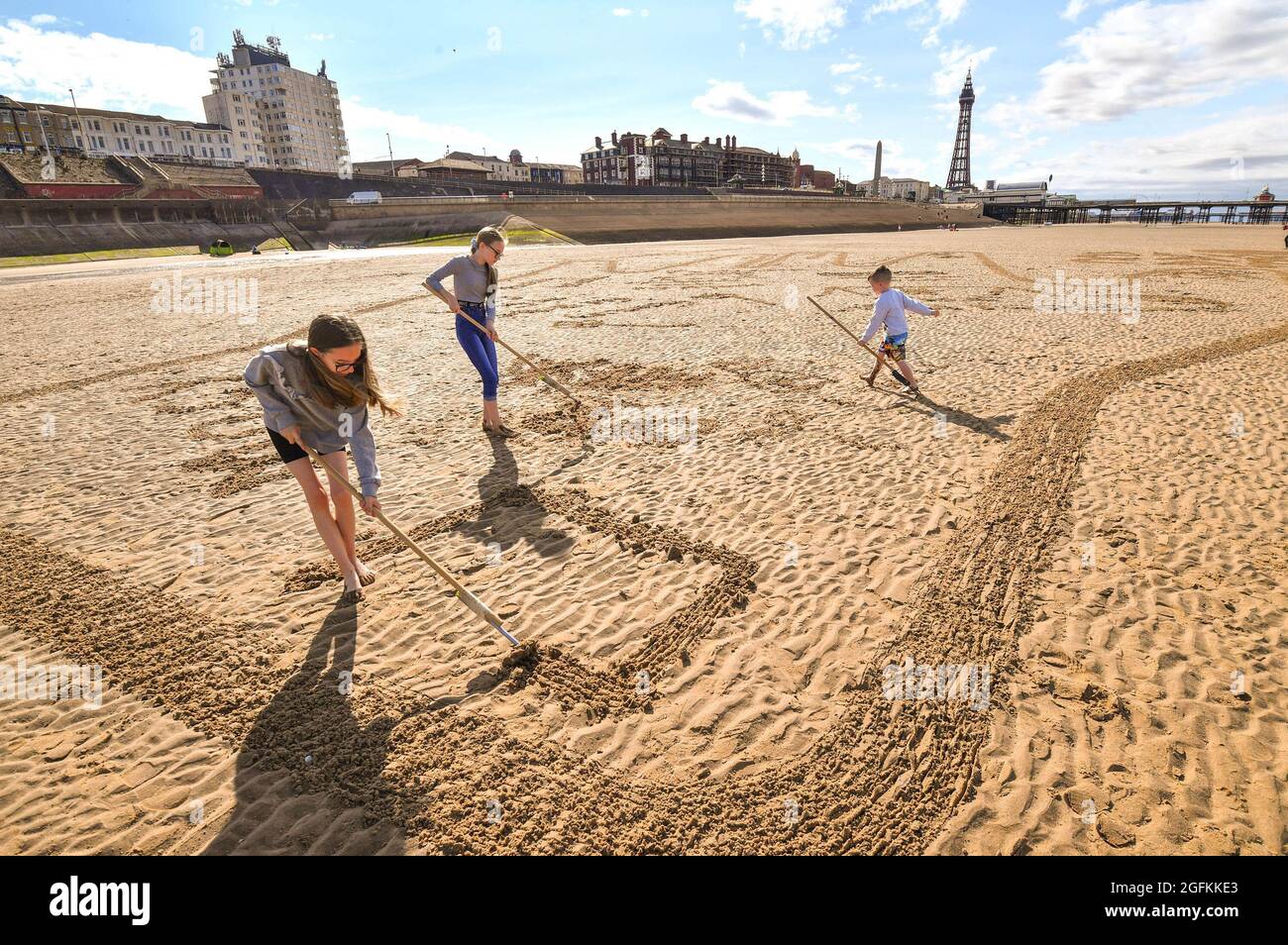 EDITORIAL USE ONLY (Left to right) Macy, Hollie and Marshall from Manchester draw in the sand as members of the public contribute personalised messages to their loved-ones on a 40-metre sand drawing by artists 'Sand in Your Eye', which has been created by hygiene brand Carex to encourage people to get back together safely , Blackpool. Picture date: Thursday August 26, 2021. Stock Photo