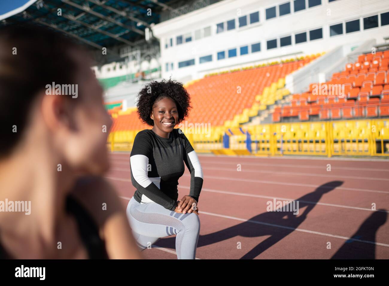 Black female runner smiling and lunging near girlfriend while warming up on track Stock Photo