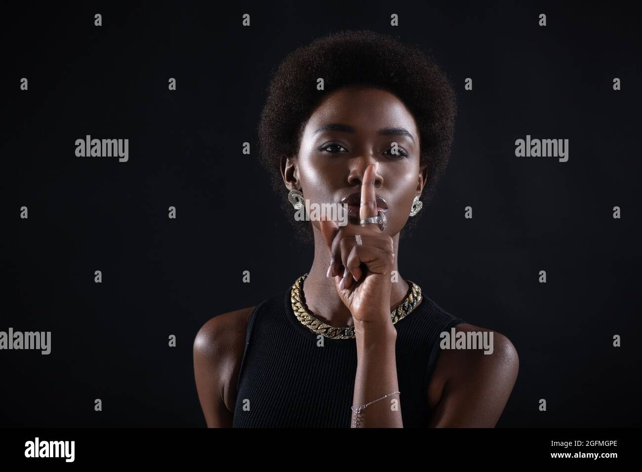 Closeup of beautiful young african american dark-skinned woman with finger on her lips showing silence gesture on black background. Stock Photo