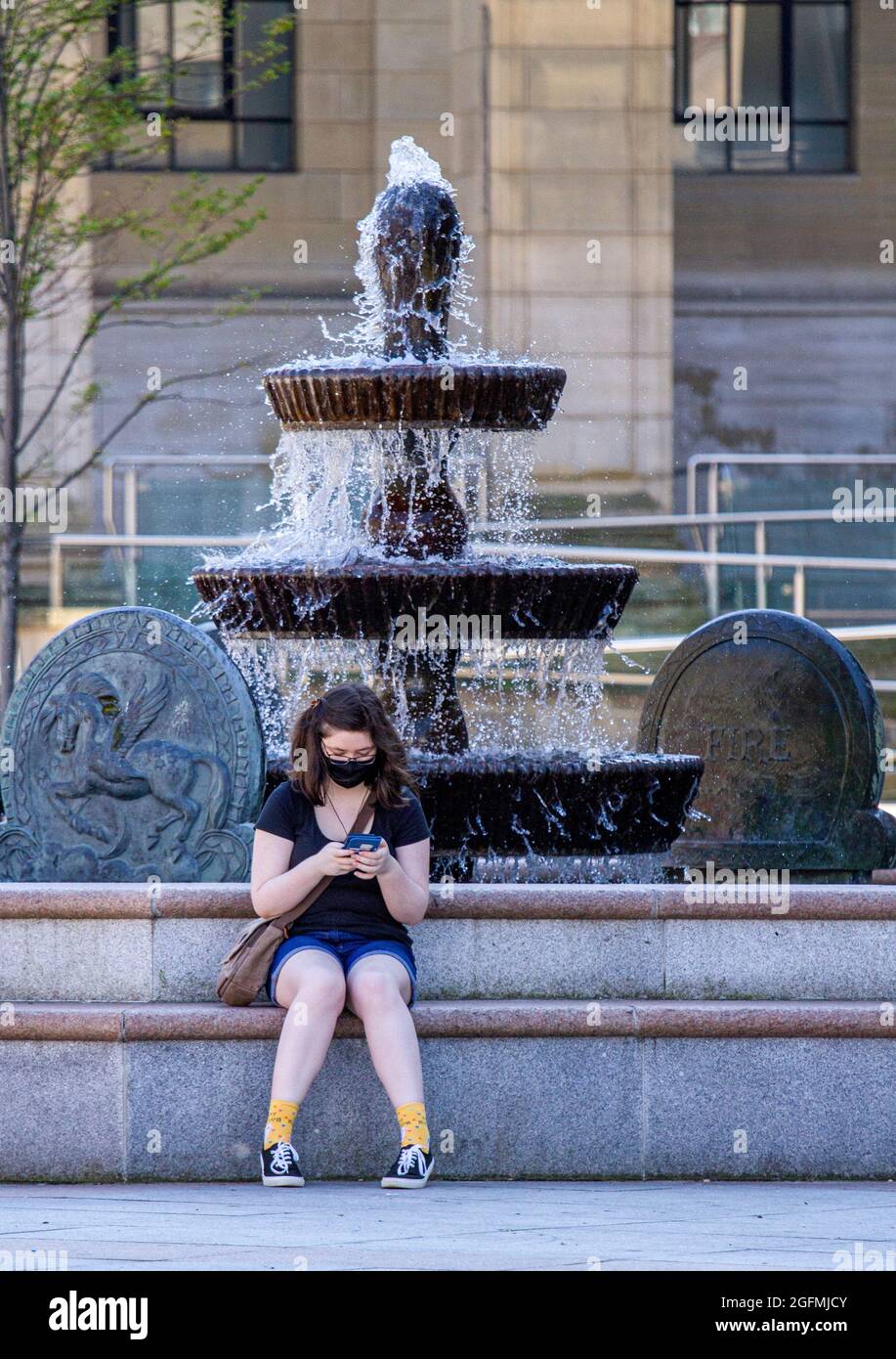 Dundee, Tayside, Scotland, UK. 26th Aug, 2021. UK Weather: A warm sunny day with a slight cool breeze across North East Scotland with temperatures reaching 20°C. A young glamorous woman wearing a facemask sitting beside the Caird Hall fountains cooling off from the warm sunshine whilst texting messages on her I Phone in Dundee city centre. Credit: Dundee Photographics/Alamy Live News Stock Photo