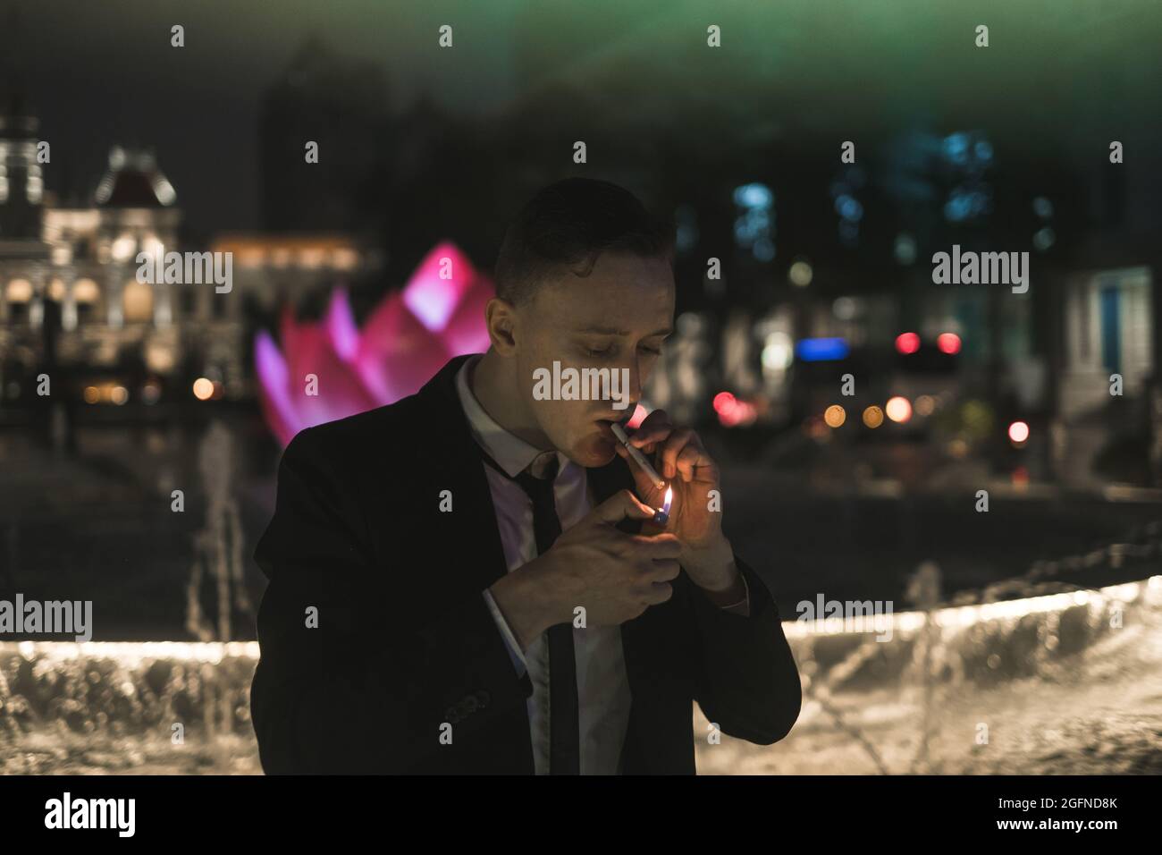 Portrait of young caucasian man in formal black suit lights a cigarette from a lighter. Night picture. Neon signs Stock Photo