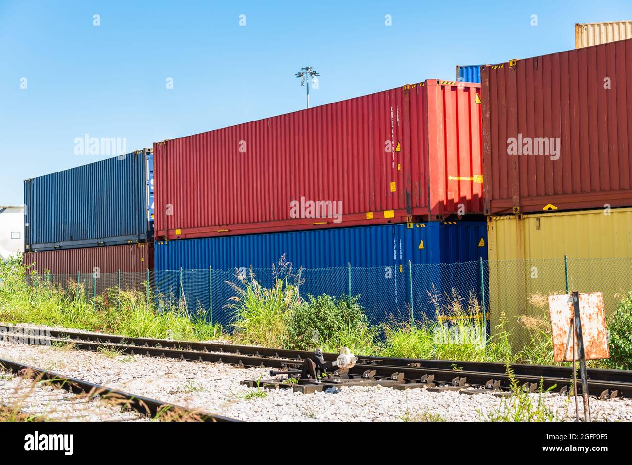 Stack of colourdul cargo containers in a fenced cargo terminal on a sunny summer day. Railway tracks are in foreground. Stock Photo