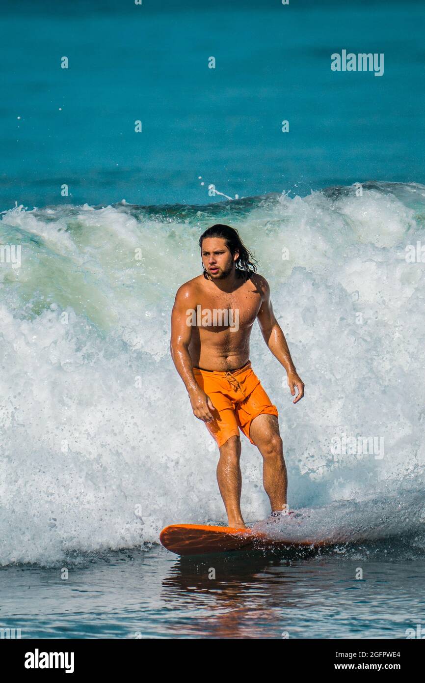 Playa Hermosa, Guanacaste, Costa Rica - 07.26.2020: A muscular long-haired latino man wearing orange shorts is surfing at the pacific Coast of Costa R Stock Photo