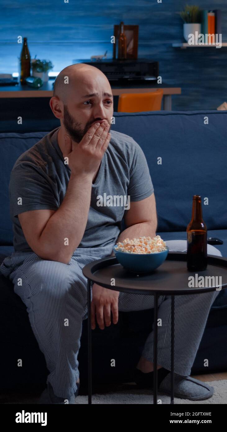 Focused man looking at drama movie, crying sitting on sofa eating ...