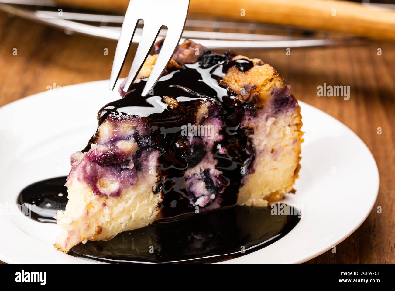 Using metal fork taking a bite from a piece of homemade blueberry and crumble cheesecake topping with chocolate in white ceramic plate on wooden table Stock Photo