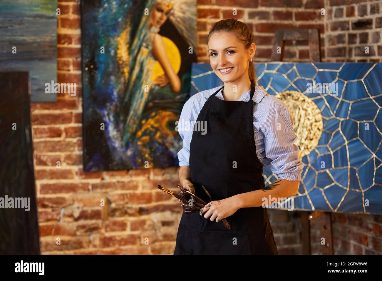 Portrait of young smiling female artist wearing apron in art studio ...