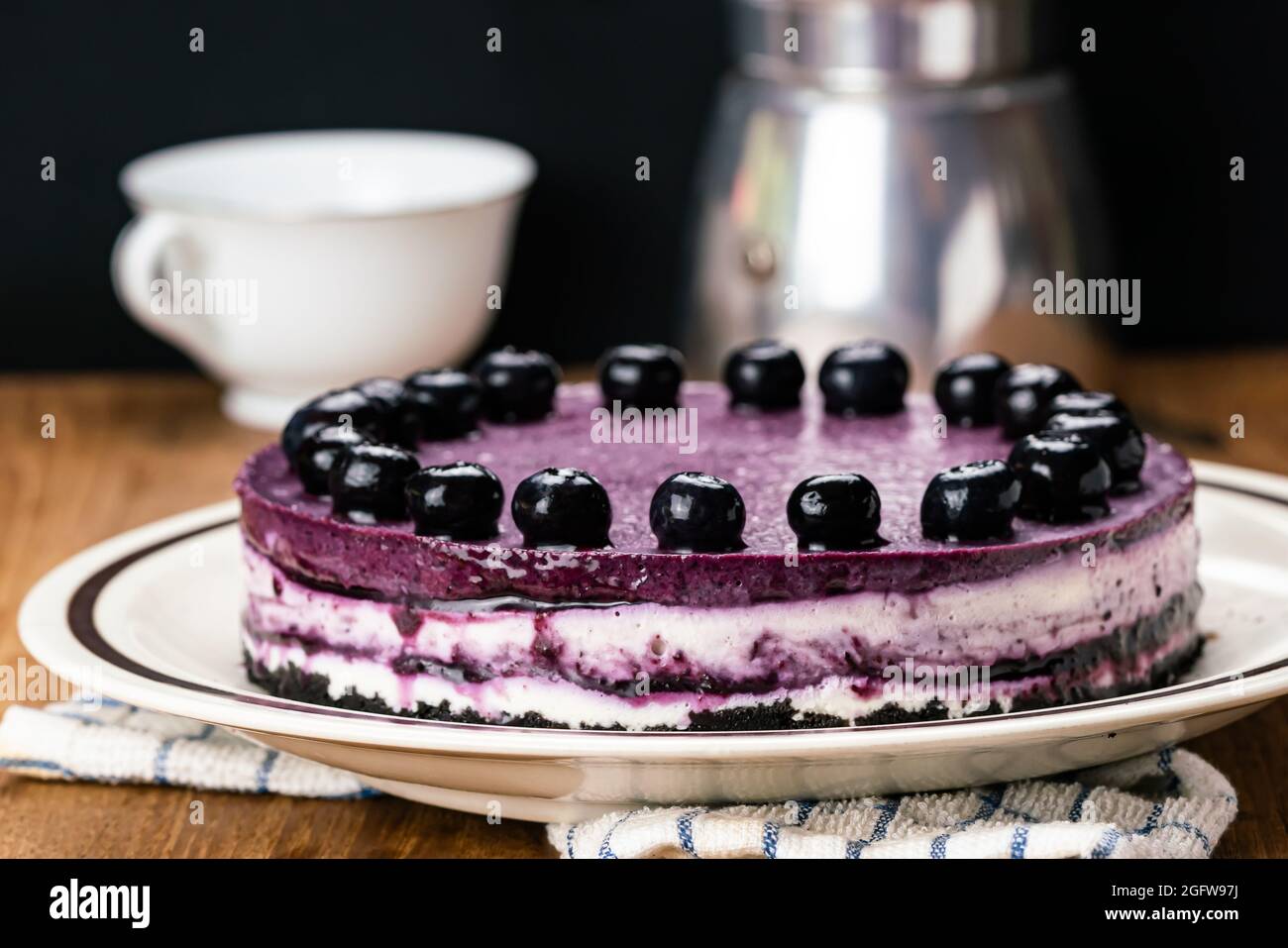Side view of delicious homemade blueberry cheesecake garnished with preserved blueberry in brown ceramic dish with moka pot coffee maker and white cer Stock Photo