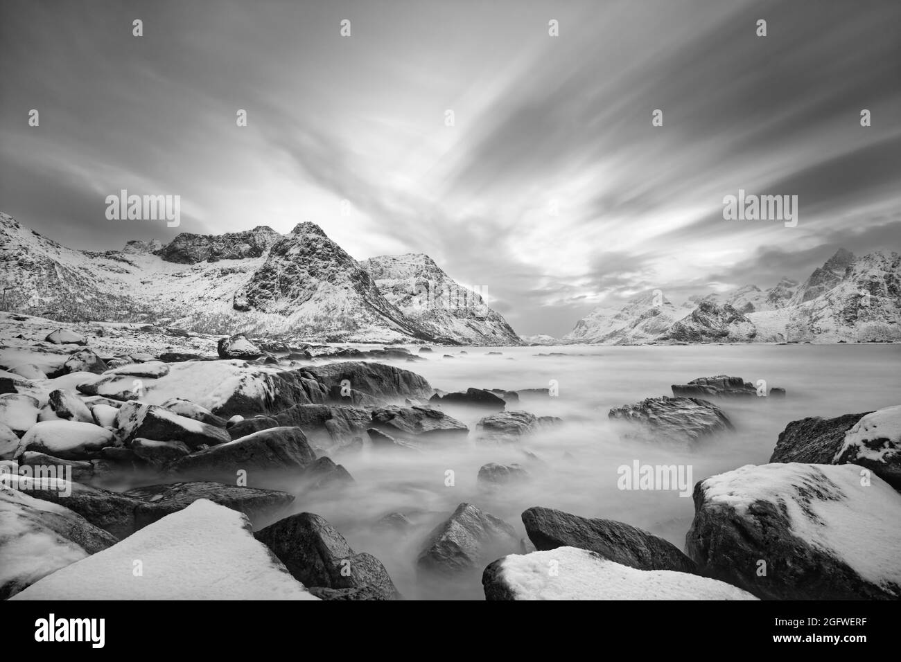 Black and white image of a coastal landscape in winter with water movement between large stones with snow on them, in the background a mountain range Stock Photo