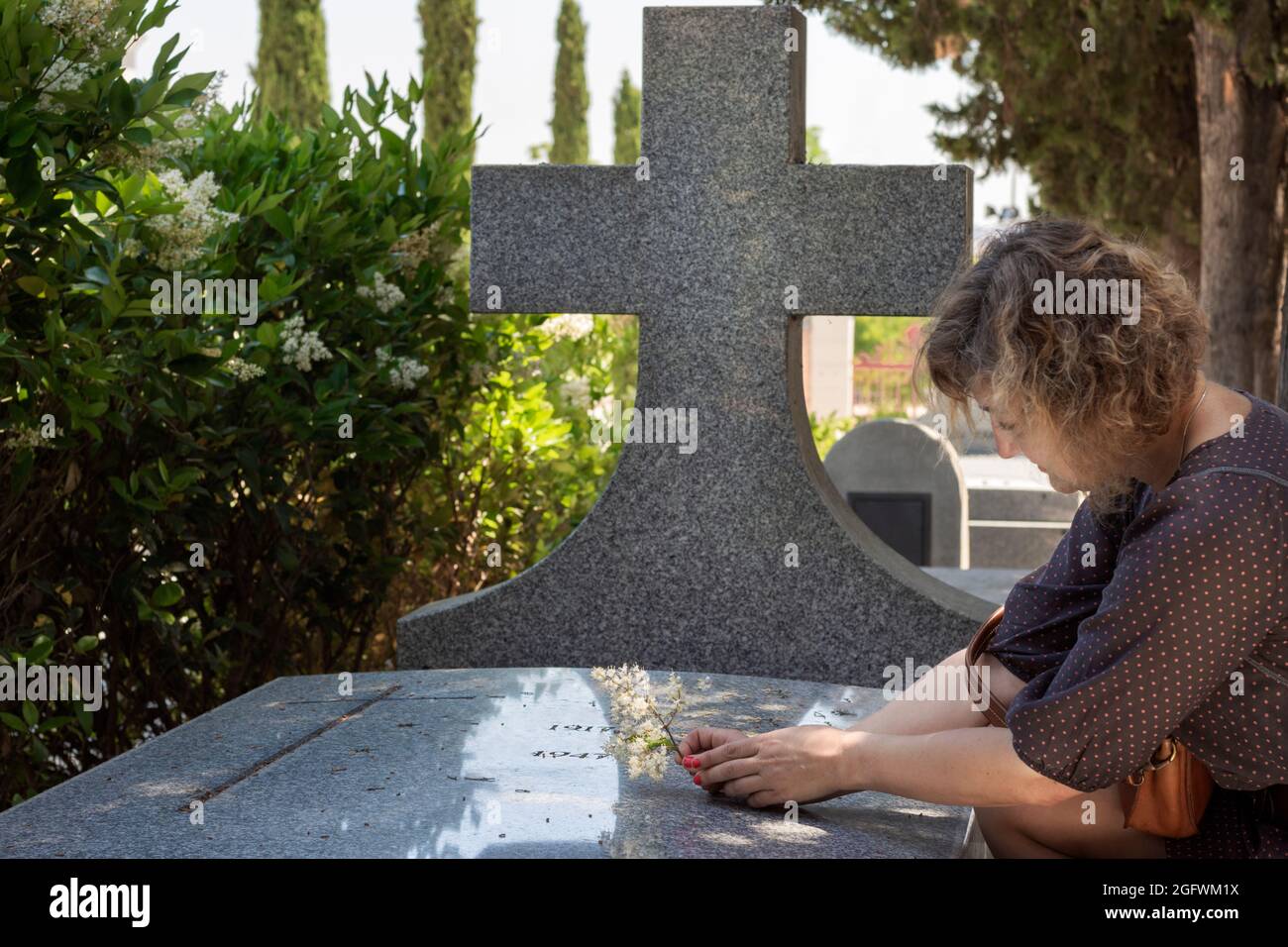 Young woman,with head bowed and sad kneeling beside a marble tomb with a large cross and a cross engraved, leaving a sprig of flowers on a grave. In t Stock Photo