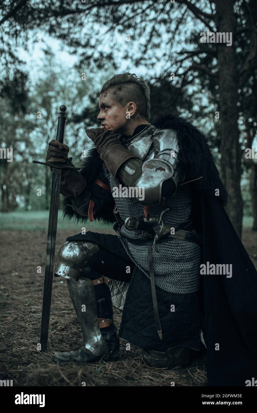 Medieval warrior knight in chain mail armour and black cape standing on one  knee among forest with his sword Stock Photo - Alamy