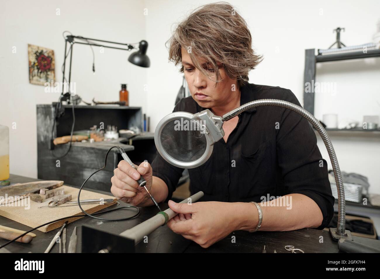 Concentrated female jeweler sitting in workshop with black furniture and soldering pieces of jewelry together under magnifying glass Stock Photo