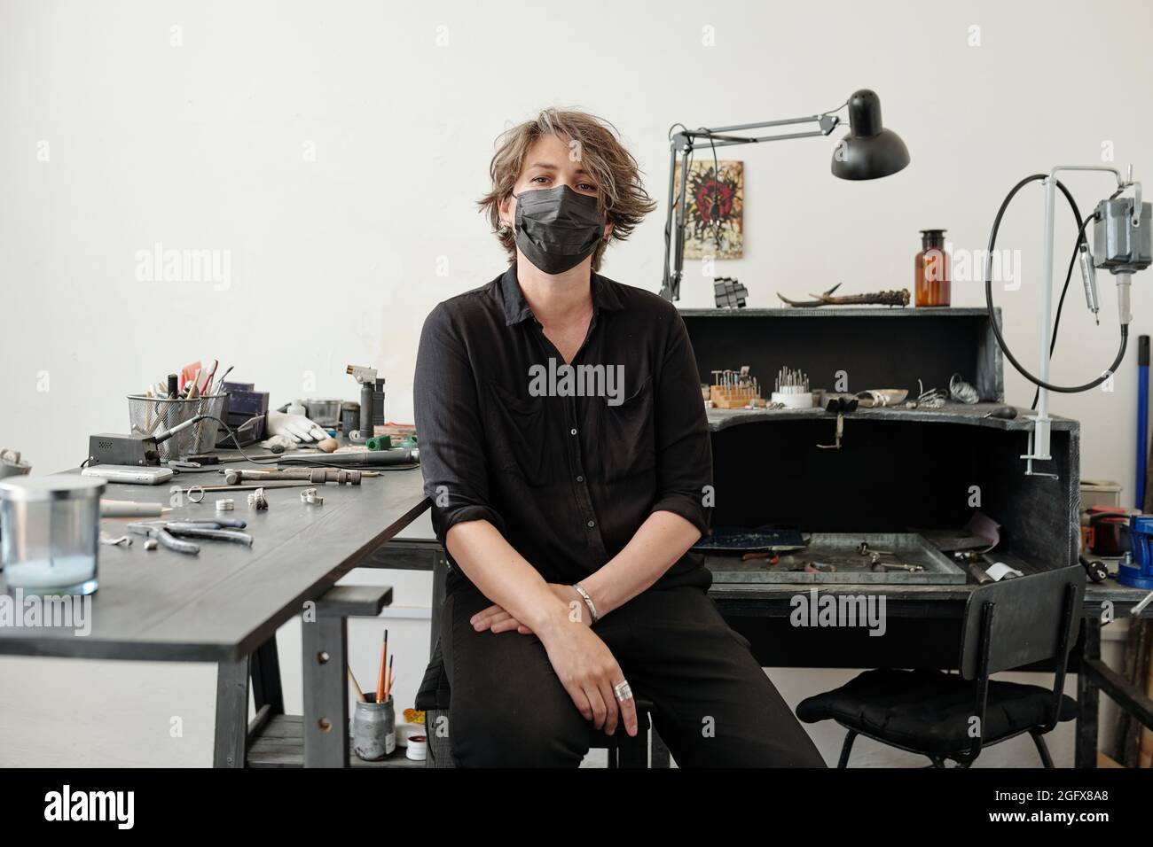 Portrait of female jeweler with short hair wearing mask sitting on stool against work bench in workshop Stock Photo