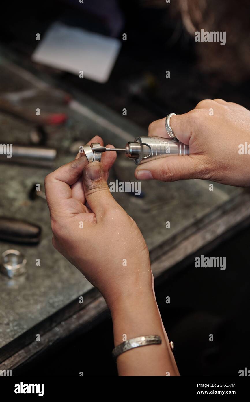 Close-up of female jeweler with dirty hands cleaning silver ring with polisher in dark workshop Stock Photo