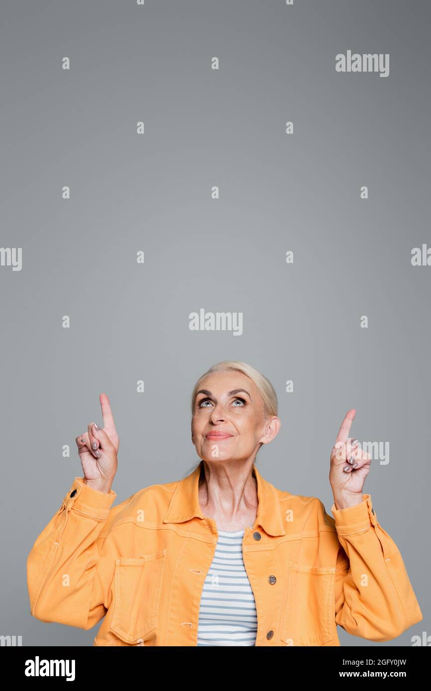 smiling elderly woman in orange jacket looking up and pointing with fingers isolated on grey Stock Photo