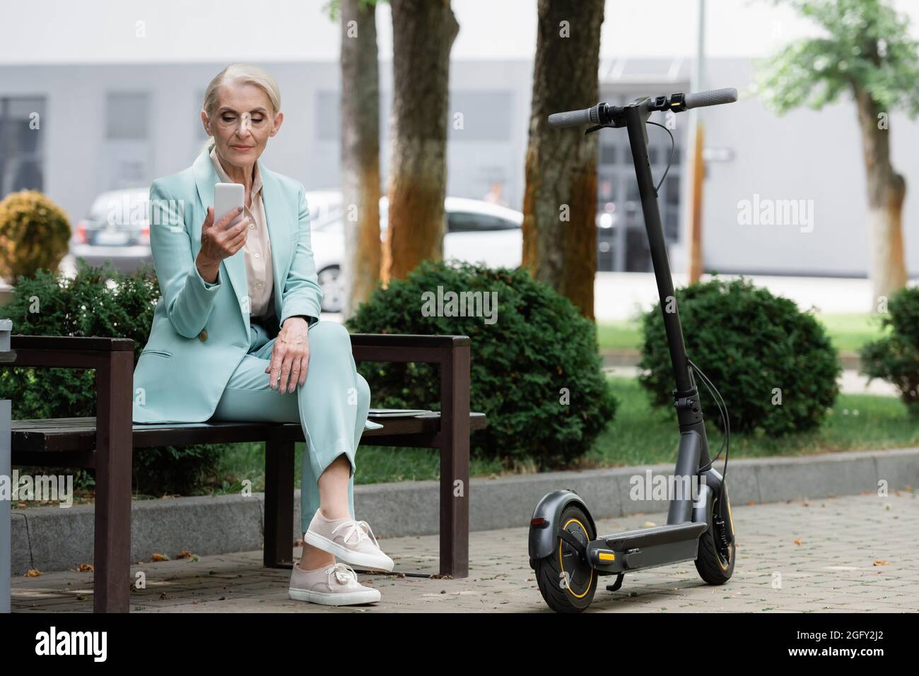 senior businesswoman using smartphone while sitting on bench near electric kick scooter Stock Photo