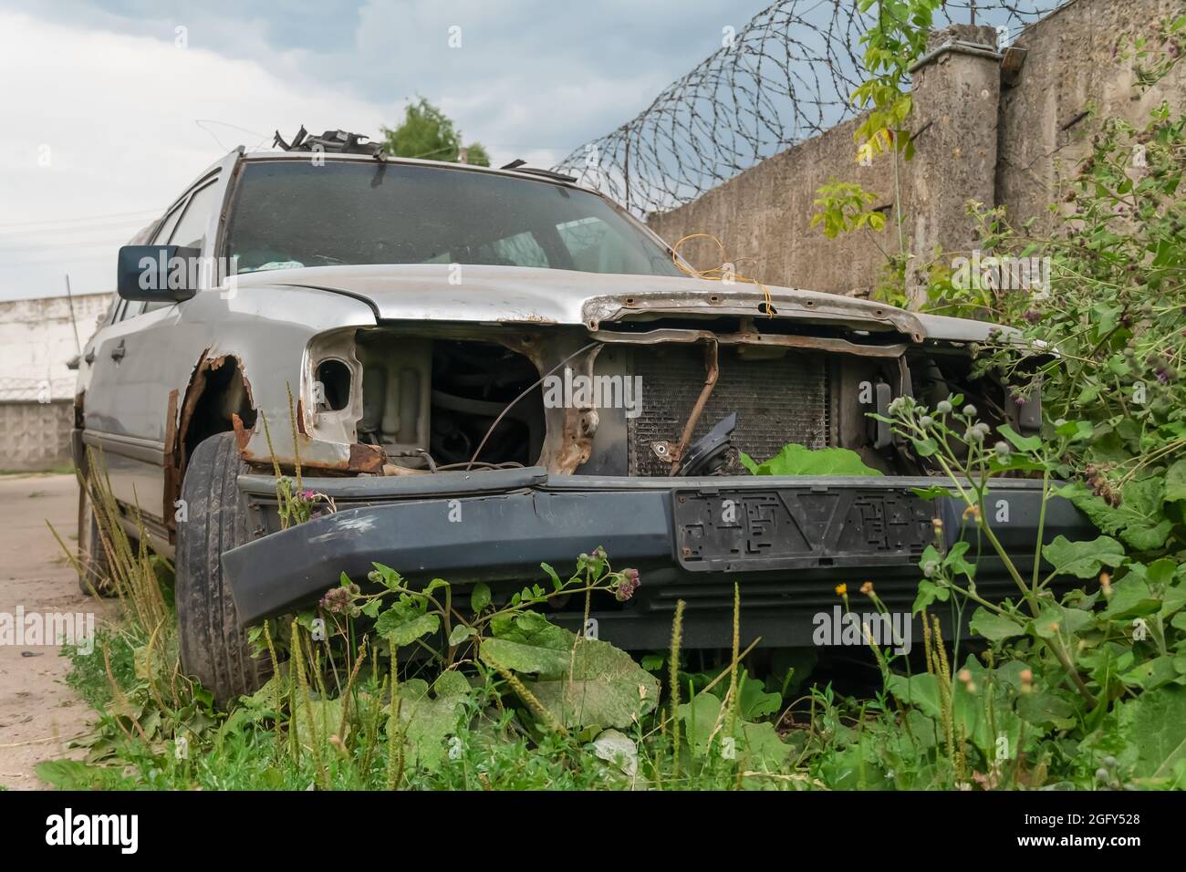 Old rusty gray car without headlights near a concrete fence with barbed