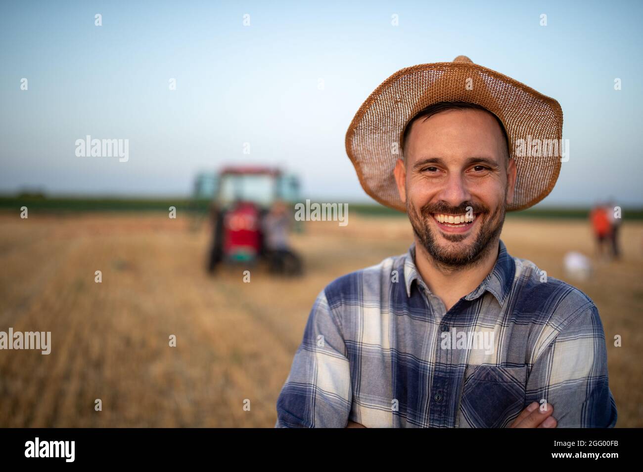 Portrait of satisfied handsome farmer with straw hat on head in field during harvest with tractor in background Stock Photo