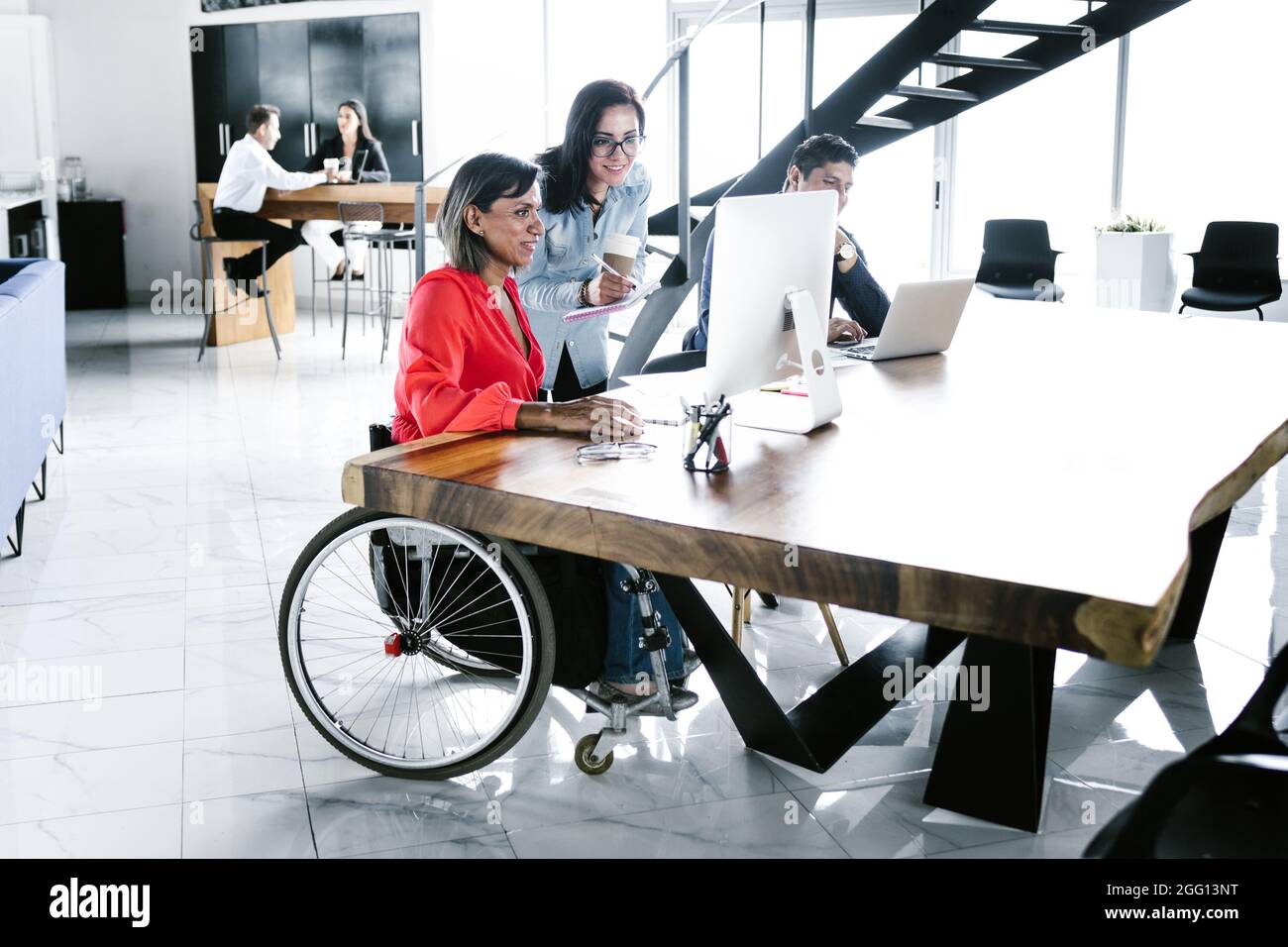 Hispanic transgender woman sitting in wheelchair and coworker looking at computer at workplace in Latin America Stock Photo