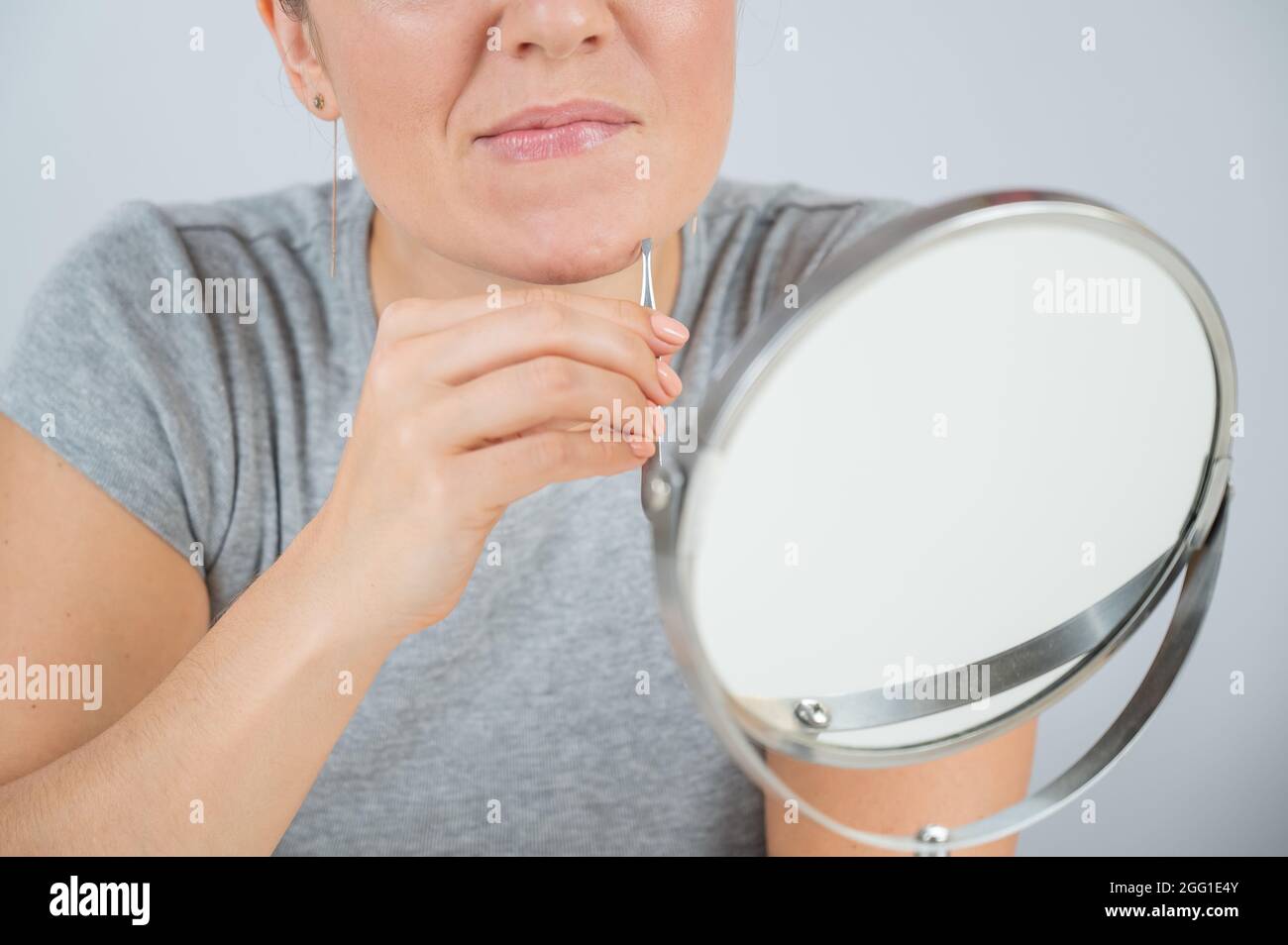Caucasian woman looks in the mirror and removes hair on her chin herself with the help of tweezers. Hirsutism. Stock Photo