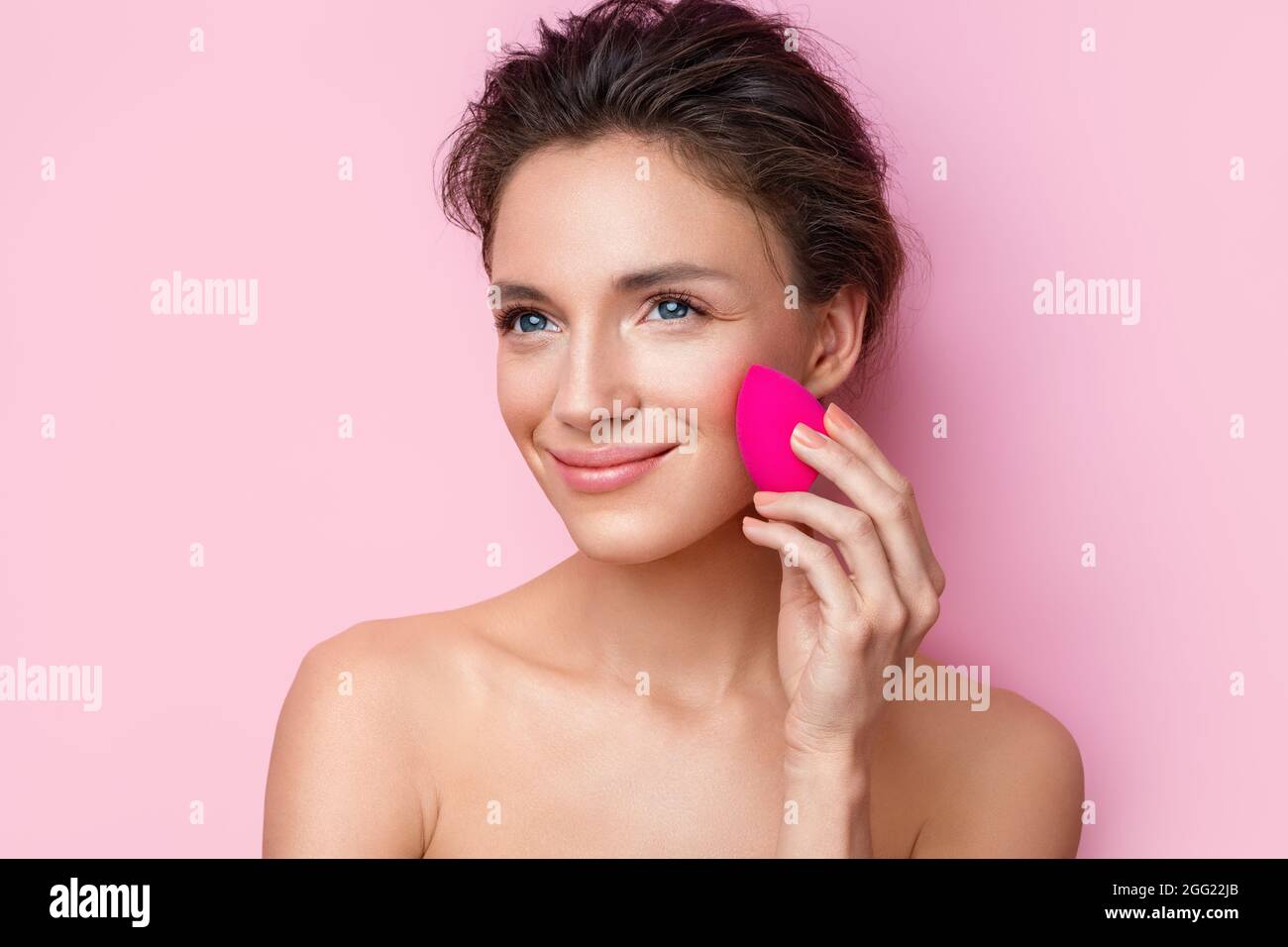Woman applying foundation using cosmetic sponge, beauty blender. Photo of woman with perfect makeup on pink background. Beauty concept Stock Photo