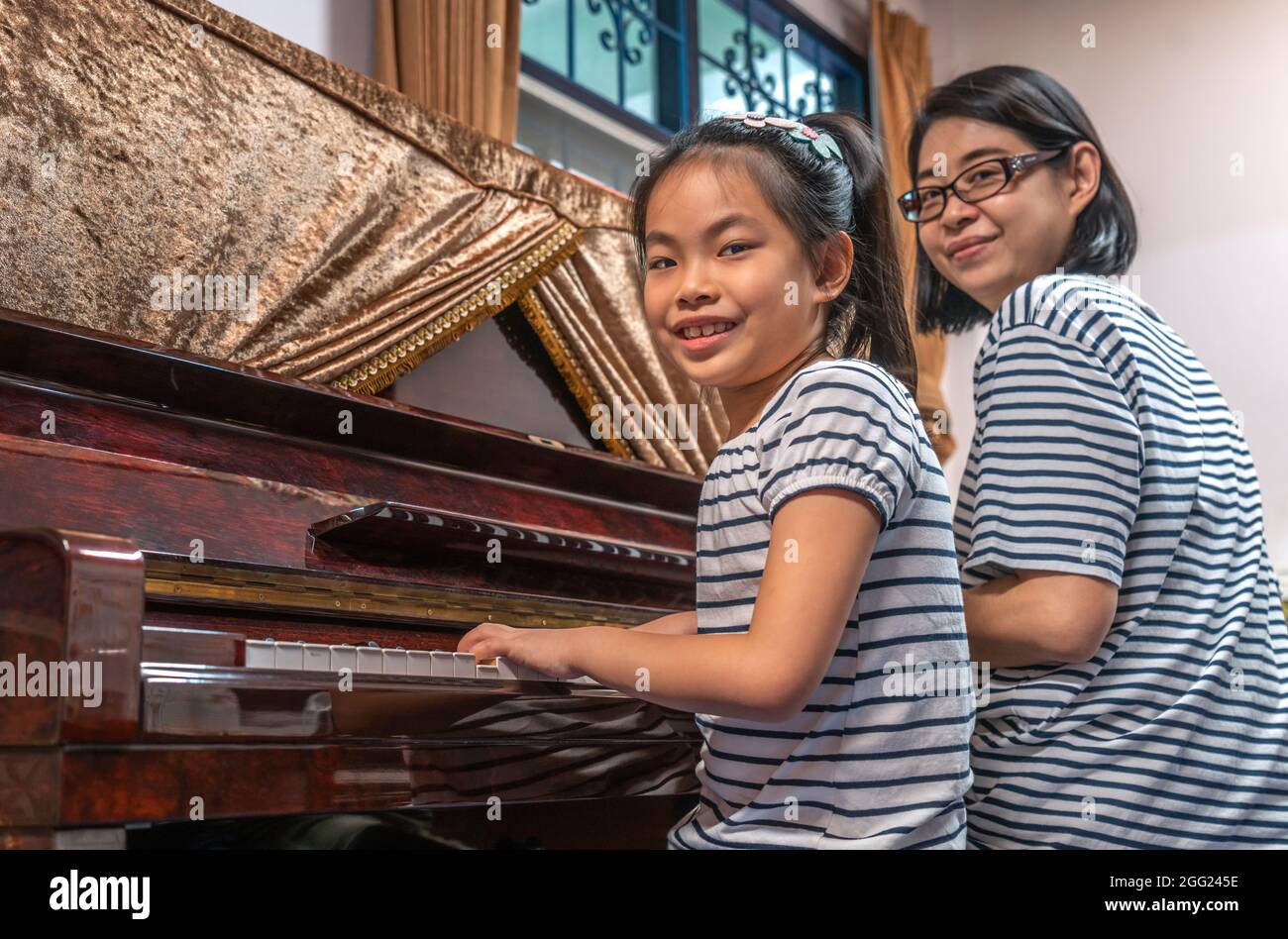 Asian cute child girl practicing pianon lesson white her mother at home, playing upright piano, looking to camera and smile. Stock Photo