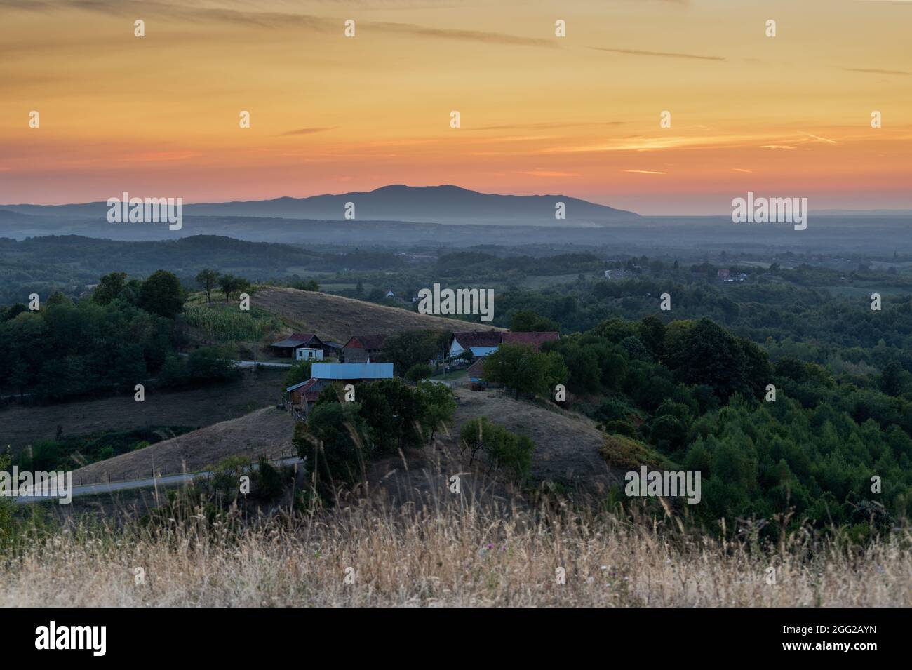 Hilly countryside landscape with farmhouse, meadows, forests and distant mountain in haze, beautiful vivid color in sky at dusk Stock Photo