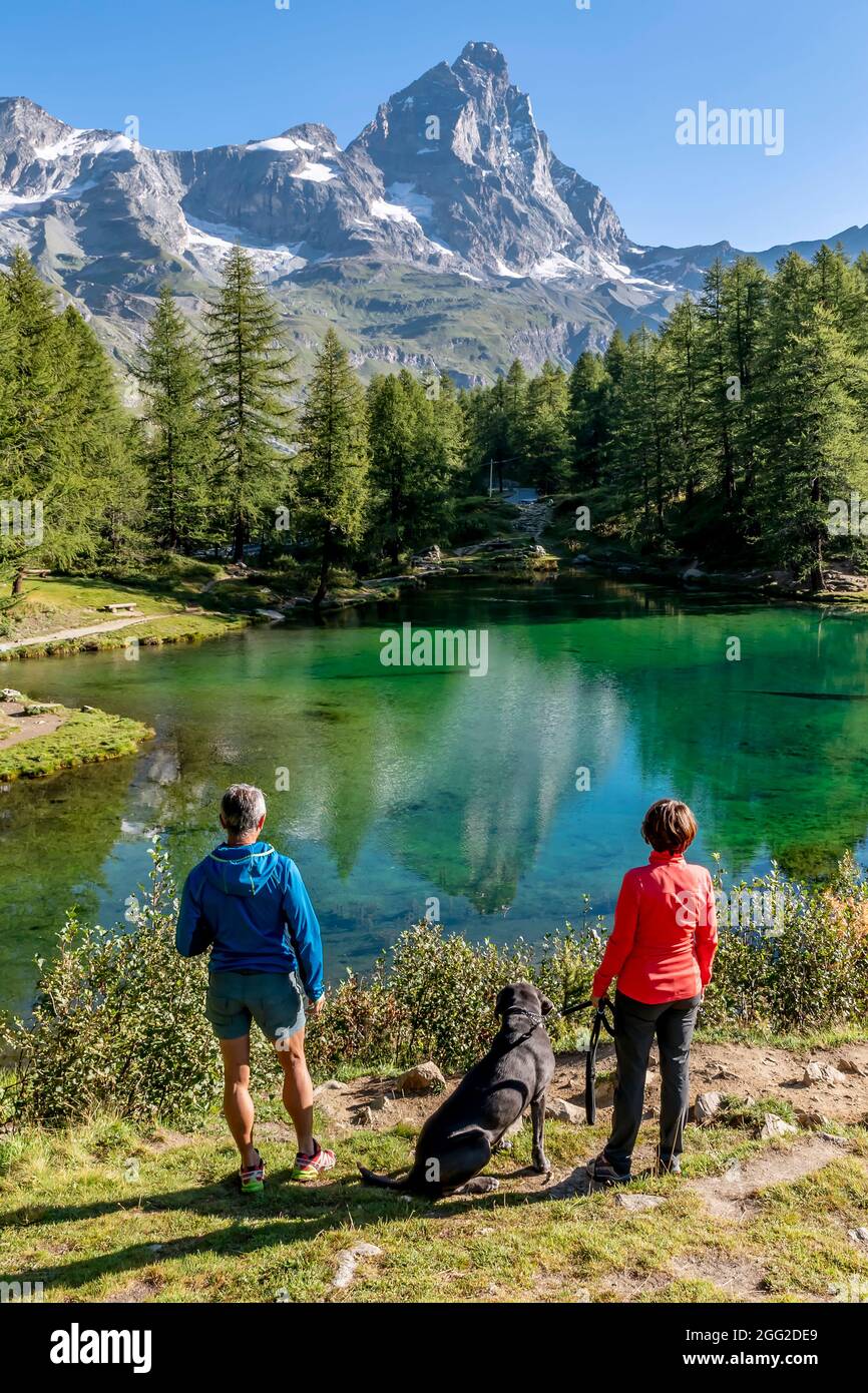 A couple with a dog admires the beautiful landscape of Lago Blu or Layet, which mirrors Mount Cervino, Aosta Valley, Italy Stock Photo