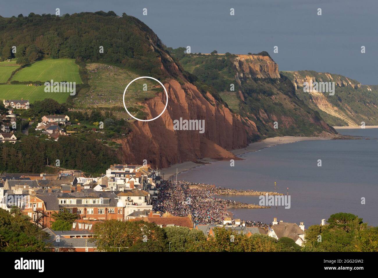 Sidmouth, Devon, UK. 27th Aug 2021.  Members of the public stand dangerously close to the edge of crumbling cliffs whilst watching the RAF Red Arrows perform a colourful display over Sidmouth, Devon. The cliffs at Sidmouth have collapsed in this location around half a dozen times in the past 4-6 weeks. Note: White circle indicates location where public were viewing the display. Credit: Ian Williams/Alamy Live News Stock Photo