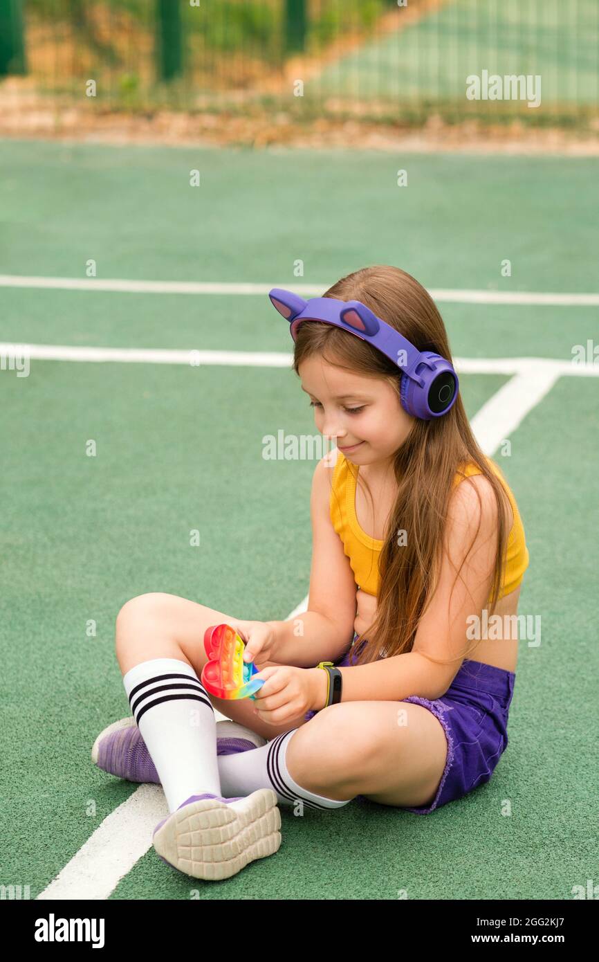 School girl is playing with modern form heart popit toy. Relaxing on sports ground feel no stress free. Silicone sensory toy antistress in hands. Simple dimple. Stock Photo