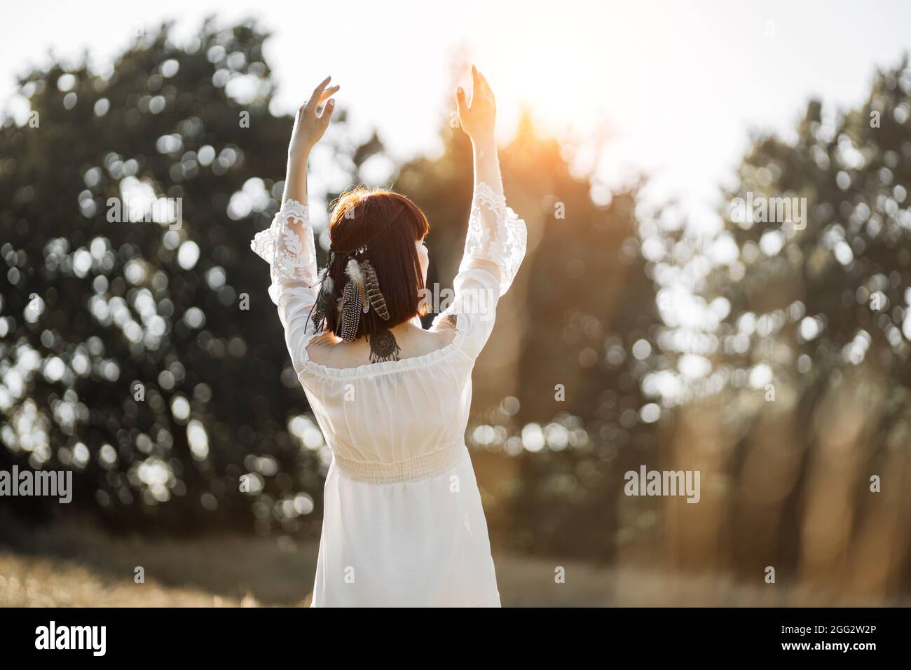 Back rear portrait of charming young native american woman in white boho dress and with feathers in hair, posing face to summer sunset, raising her ar Stock Photo